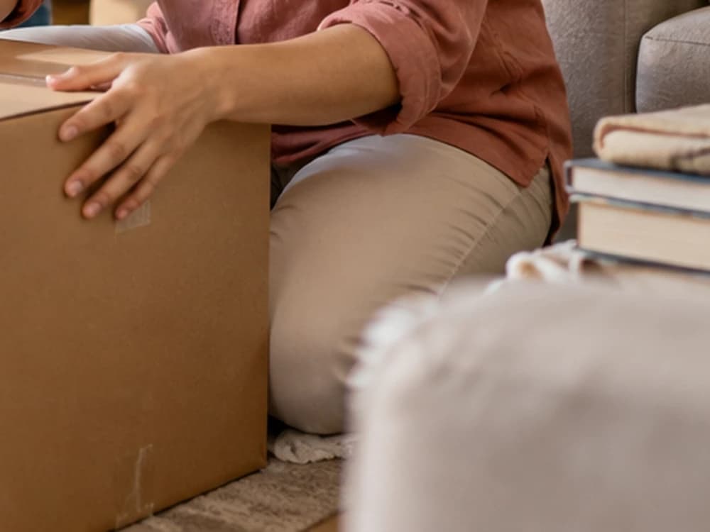 A family member handling a moving box beside books and bedding